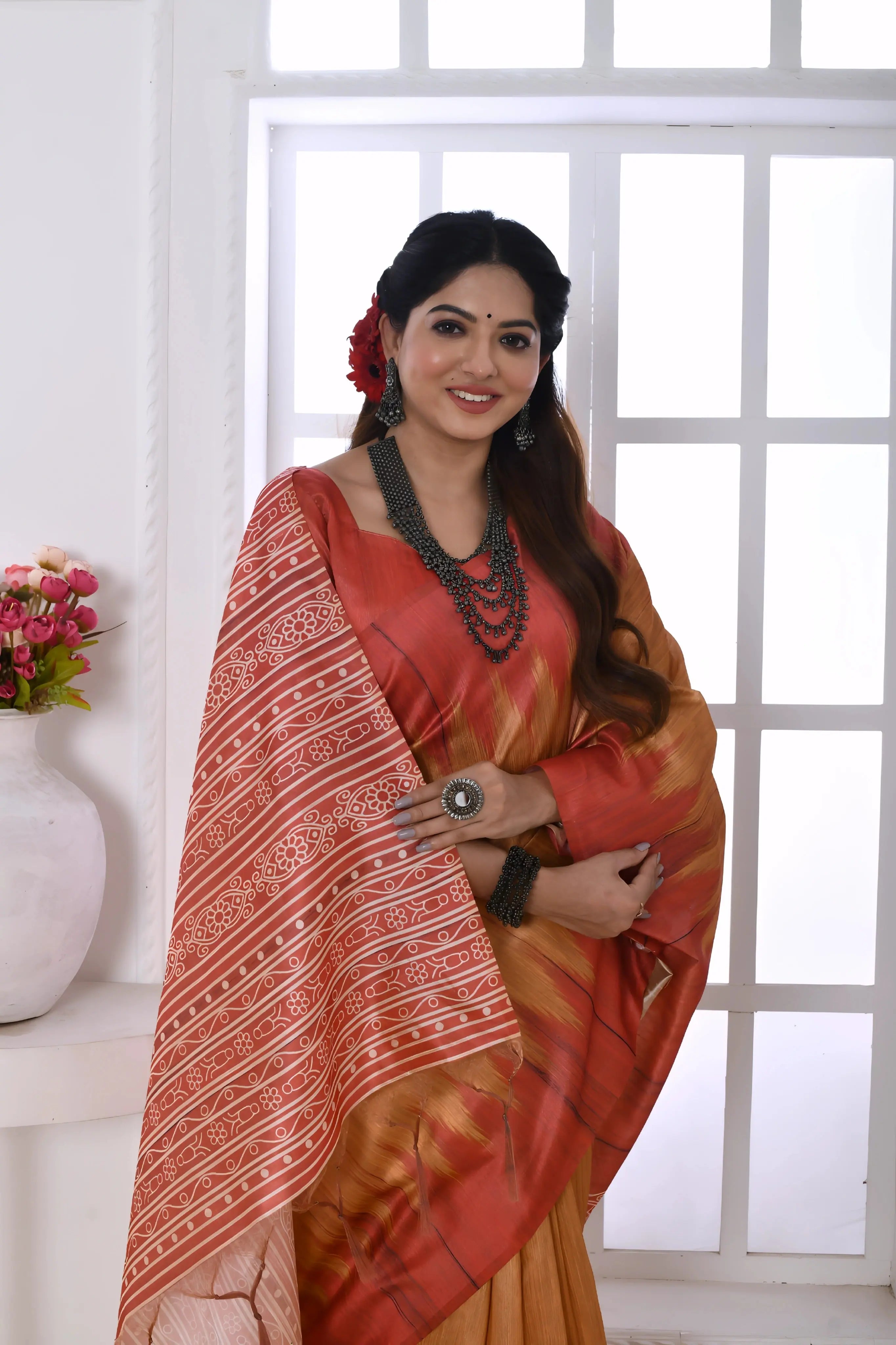 A woman models a graceful Copper Orange Soft Tussar Silk Saree. The saree features a zigzag pattern where the body meets the pallu, and the pallu has a wide panel of dense white and red geometric tribal-style print. She wears a coral blouse and a chunky dark beaded necklace, posing indoors by a large window.